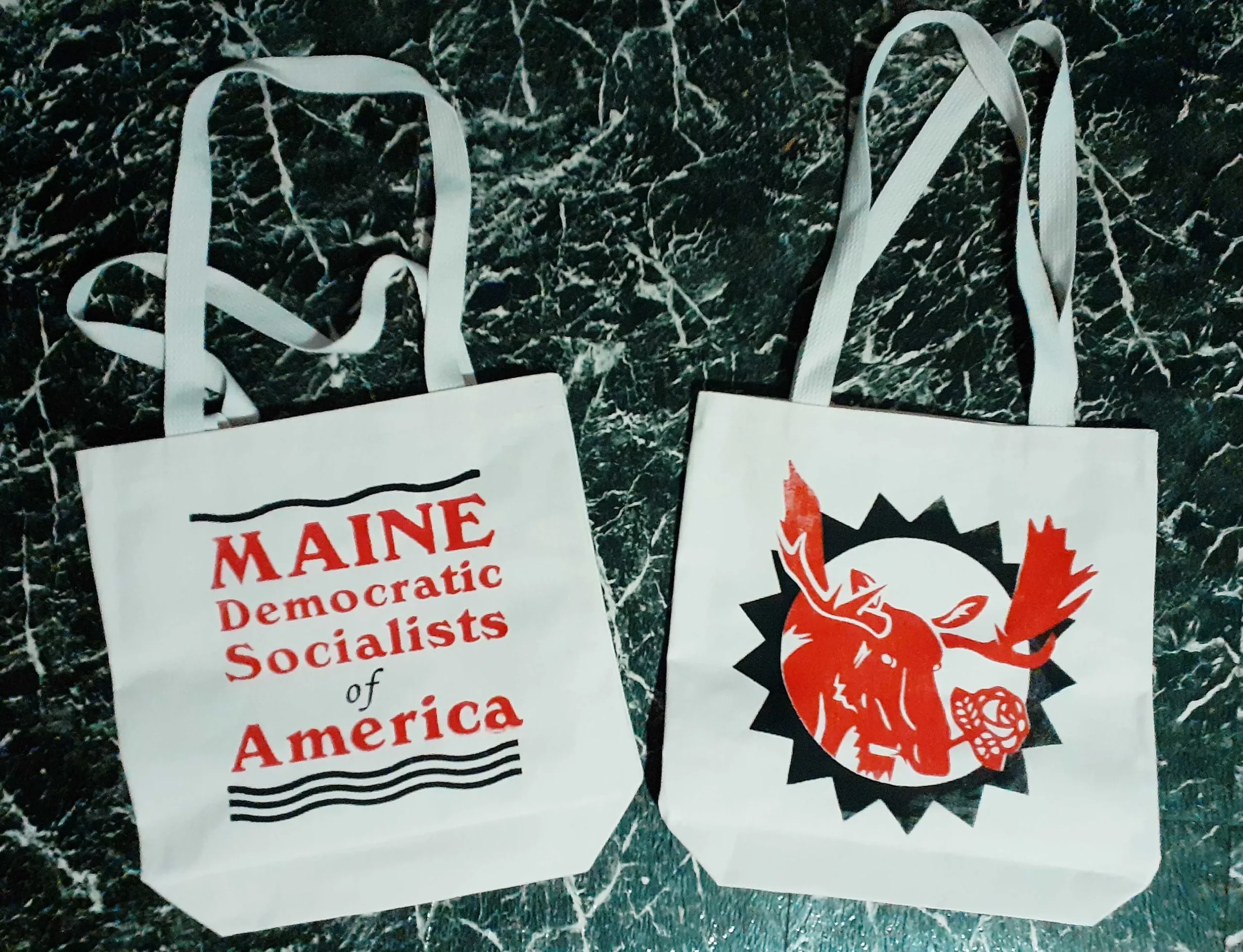 Exclusive Maine DSA Merch screen printed by a local comrade on canvas totes made in New Hampshire. A moose holding a rose in its mouth on one side and Maine Democratic Socialists of America on the other with wavy lines above and below the text.