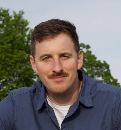 a white man with short hair, a trim mustache, and a simple navy blue button up over a white undershirt poses outside with a sincere expression