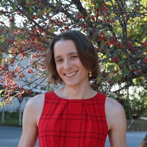 a white woman in a red dress a gold floral earrings stands outside with a wide smile in front of a crabapple tree